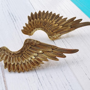 Op de afbeelding: Twee gouden metalen engelenvleugels met een gedetailleerd verenontwerp. De vleugels zijn licht gebogen en hebben een getextureerd oppervlak.