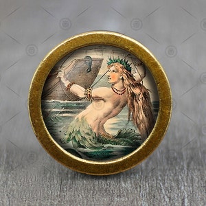 May include: A brass knob with a round, gold-colored frame and a circular image of a mermaid with long, flowing hair, a green tail, and a crown of leaves. The mermaid is holding a rope and is surrounded by water.