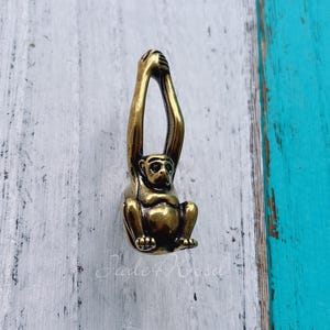 May include: A gold-toned monkey-shaped cabinet knob or pull. The monkey is hanging upside down with its arms raised above its head. The monkey has a detailed face and body.