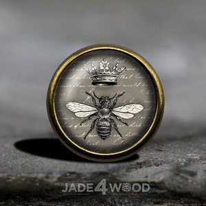 May include: A round, gold-toned metal button with a glass top featuring a black and white illustration of a bee with a crown above it. The background is a gray and white pattern of script.
