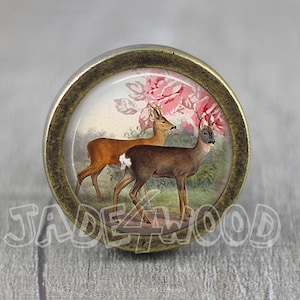 Op de afbeelding: Een ronde, antieke goudkleurige knop met een glazen inzetstuk met een vintage illustratie van twee herten in een bosrijke omgeving met roze bloemen.