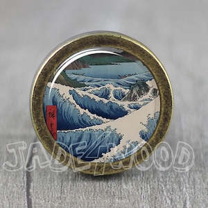 Op de afbeelding: Een ronde, messingkleurige kastknop met een glazen inzetstuk met een Japanse houtsnede van een grote golf die tegen een rotsachtige kust breekt. De golf is blauw en wit, en de kust is bruin en groen. De print is omgeven door een messingkleurige lijst.
