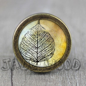 Puede incluir: Un pomo redondo de color latón con un inserto de vidrio que presenta una imagen en blanco y negro de una hoja sobre un fondo amarillo y blanco.