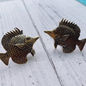 May include: Two gold-colored fish-shaped decorative drawer pulls. The fish have a detailed, scaly design and are facing opposite directions.