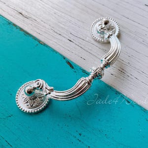 May include: Silver drawer pull with an ornate design. The pull has a curved shape and a decorative knob on each end.