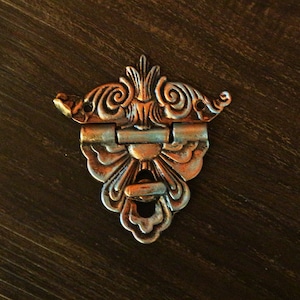 Puede incluir: Pestillo de metal plateado adornado con un diseño floral decorativo. El pestillo tiene una sección con bisagras y un pequeño ojo de cerradura.