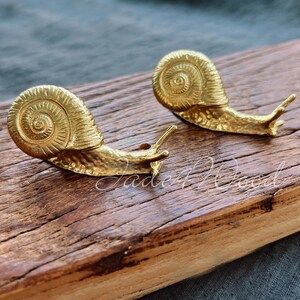 May include: Two gold snail sculptures on a wooden surface. The snails are detailed with a spiral shell and a textured body.