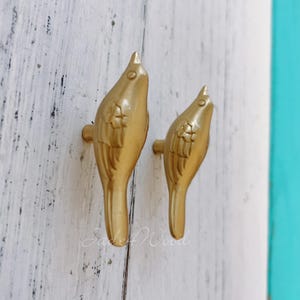 May include: Two gold-colored bird-shaped cabinet knobs. The knobs are mounted on a white, distressed wooden surface. The birds are facing upwards with detailed wing and tail features. The knobs are likely for a drawer or cabinet.