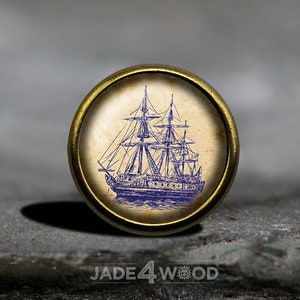May include: A gold-toned metal button featuring a blue and white illustration of a sailing ship on a vintage-style background.