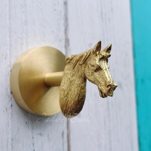 May include: A gold-coloured cabinet knob in the shape of a horse's head. The knob has a detailed sculpture of a horse's head, mounted on a circular base. The knob is attached to a white wooden surface.