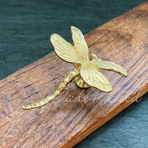 May include: A gold-toned dragonfly door knocker with intricate details. The knocker is mounted on a piece of rustic wood.