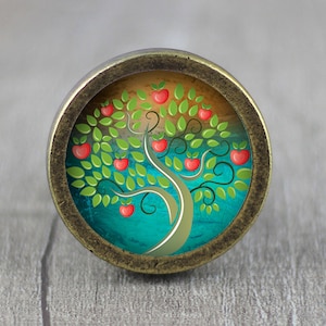 May include: A round, bronze-coloured knob with a colourful image of a stylised tree with red apples and green leaves. The tree is set against a blue and yellow background.