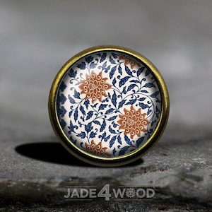 Puede incluir: Un pomo redondo de metal dorado con fondo blanco y un patrón floral azul y naranja. El pomo tiene un borde elevado.