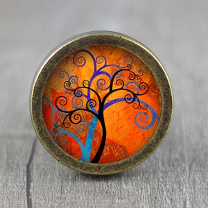 May include: A round, bronze-colored cabinet knob with a colorful, abstract tree design. The tree has a black trunk and branches with blue and purple swirls on an orange background.