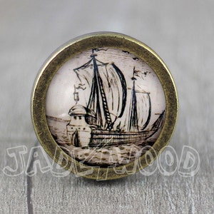 May include: A round, brass-colored knob with a vintage-style image of a sailing ship on a white background. The ship has a brown hull and black sails.