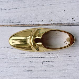 Pode incluir: Um puxador de armário dourado, em forma oval e com um design curvo. O puxador tem uma superfície lisa e refletora e é montado numa superfície de madeira branca. O puxador tem uma pequena abertura no centro.