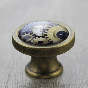 Könnte beinhalten: Ein Messing-Schrankknauf mit einer runden, gewölbten Oberseite, die ein Steampunk-Design mit Zahnrädern und einem Kompass zeigt. Der Knauf hat eine strukturierte Oberfläche.
