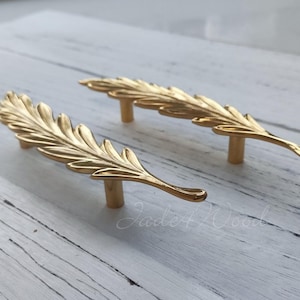 May include: Two gold leaf-shaped cabinet pulls with a detailed design. The pulls are on a white wooden surface.