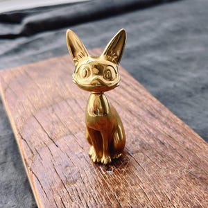 Puede incluir: Una figurita de metal dorada de un gato con grandes ojos expresivos. El gato está sentado derecho con la cola enroscada alrededor de su cuerpo.