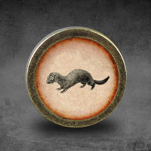 May include: A round, brass-colored button with a raised, orange-brown rim. The button features a black and white image of a weasel or ferret running to the right.