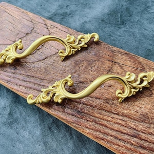 Op de afbeelding: Twee sierlijke, goudkleurige kastgrepen met een gebogen ontwerp en decoratieve krullen. De handgrepen worden getoond op een verweerd, bruin houten oppervlak, wat een vintage esthetiek creëert. De handgrepen zijn waarschijnlijk van metaal.