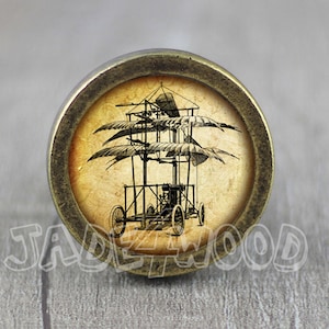 May include: A round, brass-colored knob with a vintage-style image of a flying machine with wings and wheels. The image is on a cream-colored background.