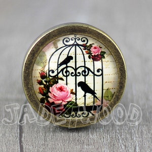 Puede incluir: Un pomo redondo de color dorado antiguo con un inserto de vidrio que presenta una jaula de pájaros negra con dos pájaros negros y rosas rosas. El fondo es un diseño estampado en blanco y negro.