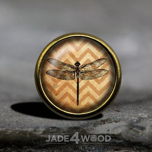 May include: A gold-toned metal button with a glass top featuring a black dragonfly silhouette on a chevron patterned background.