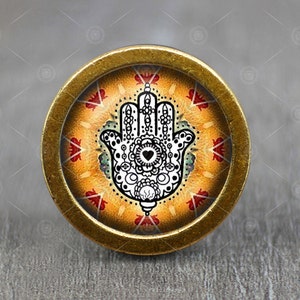 以下が含まれることがあります： アンティーク真鍮色の丸いキャビネットノブ。 ハムサハンドのデザインが特徴です。 ハムサハンドは白と黒のディテールで、暖かくオレンジと黄色の背景に赤いアクセントが施されています。 家具の装飾要素です。