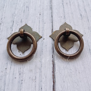 Op de afbeelding: Twee antieke bronzen kastgrepen met ringgrepen. Elke greep heeft een decoratieve achterplaat met een bloemmotief. De ronde ringgrepen zijn bevestigd aan de achterplaten. De grepen staan op een wit houten oppervlak.