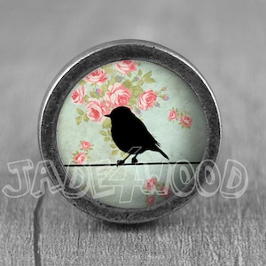 May include: Round, silver-toned cabinet knob featuring a black bird silhouette perched on a wire. The knob's center has a light blue background with pink roses. The text "JADE4WOOD" is visible.