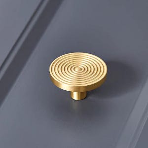 Puede incluir: Un pomo de armario dorado con un diseño circular acanalado. El pomo tiene un vástago corto y cilíndrico y una parte superior plana y redonda con círculos concéntricos. El fondo es gris apagado.
