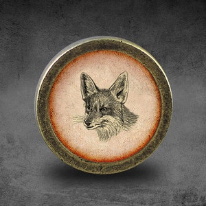 May include: A round, brass-coloured button with a raised, orange-brown rim. The centre of the button features a black and white illustration of a fox's head.