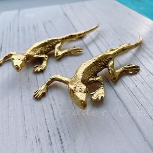 Könnte beinhalten: Zwei goldfarbene Eidechsenfiguren auf einer weißen Holzoberfläche. Die Eidechsen sind in einer kriechenden Position mit eingerollten Schwänzen dargestellt.