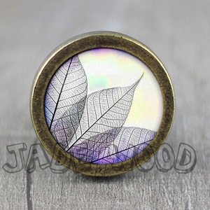 May include: Circular cabinet knob with a bronze finish. The knob showcases a close-up of translucent leaves in shades of purple and grey against a white background. A decorative element for furniture.