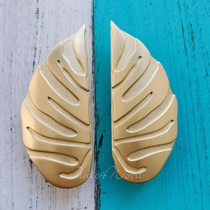 Puede incluir: Dos tiradores dorados en forma de hoja. Cada tirador tiene una superficie texturizada con una serie de líneas curvas, que se asemejan a las venas de una hoja. Los tiradores se muestran sobre un fondo de madera pintada de blanco y turquesa.