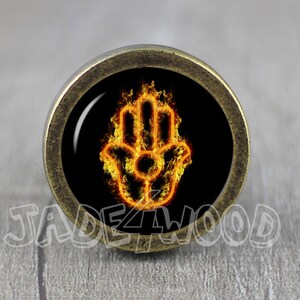 Peut inclure: Un bouton décoratif rond de couleur laiton avec un fond noir et un symbole de main de hamsa orange flamboyant.