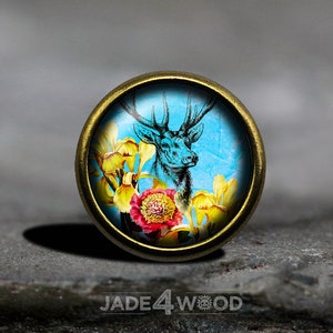 May include: A round, gold-toned metal button with a glass cabochon featuring a black and white illustration of a deer with antlers surrounded by yellow and pink flowers on a blue background.