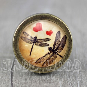 May include: A round, brass-coloured cabinet knob with a glass insert featuring two dragonflies and two red hearts on a cream background.