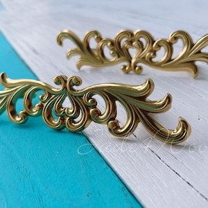 Op de afbeelding: Twee sierlijke, goudkleurige metalen ladegrepen met een decoratief scrollontwerp.