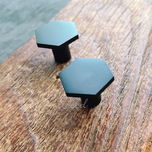 May include: Two black hexagonal cabinet knobs with a matte finish are displayed on a weathered wooden surface. The knobs have a simple, modern design, and are suitable for drawers or cabinets.