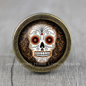 May include: A round, antique gold-toned cabinet knob with a colourful sugar skull design. The skull has orange and white details and a black spiderweb design.