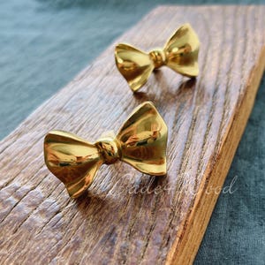 May include: Two shiny gold-coloured bow-shaped cabinet knobs. The knobs are on a wooden surface with visible grain. The knobs are likely for furniture or decorative use.