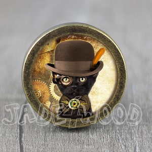 Op de afbeelding: Een zwarte kat met een bruine bolhoed met een veer, een rond steampunk-brilletje en een strik met een groen en gouden oogontwerp. De kat is geplaatst tegen een achtergrond van tandwielen en een vintage-papiertextuur.