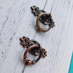 Pode incluir: Duas puxadores de gaveta de metal ornamentados com uma alça de anel. A puxador à esquerda é de cor cobre e a puxador à direita é de cor dourada.