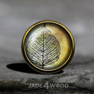 May include: A round, brass-colored pendant with a glass cabochon featuring a black and white silhouette of a leaf on a gold background.