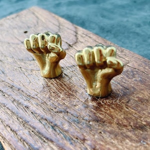 Könnte beinhalten: Zwei goldfarbene Metallfaustskulpturen auf einer Holzoberfläche. Die Fäuste sind in die Luft erhoben und symbolisieren Stärke und Einheit.