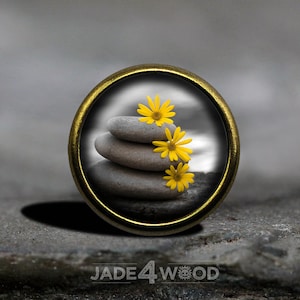 Könnte beinhalten: Ein runder Anhänger aus Messing mit einem Glaseinsatz. Der Glaseinsatz zeigt einen Stapel grauer Steine mit drei gelben Blumen darauf.