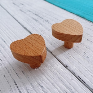 May include: Two wooden heart-shaped drawer pulls in a light brown color. The pulls are on a white wooden surface.