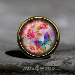 Könnte beinhalten: Ein runder, goldfarbener Metallknopf mit einem Glascabochon, der einen bunten Schmetterling auf einem Hintergrund aus rosa, roten und weißen Aquarellblättern zeigt.